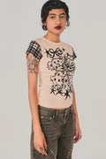 A model wearing a light pink t-shirt with a black graphic print on the front and a black-and-white plaid sleeve, paired with dark gray jeans, against a solid light gray background.