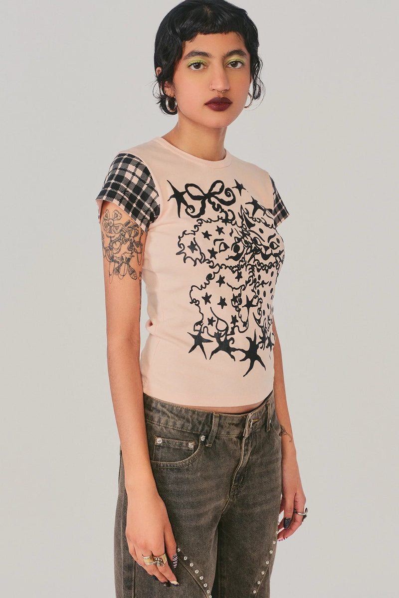 A model wearing a light pink t-shirt with a black graphic print on the front and a black-and-white plaid sleeve, paired with dark gray jeans, against a solid light gray background.