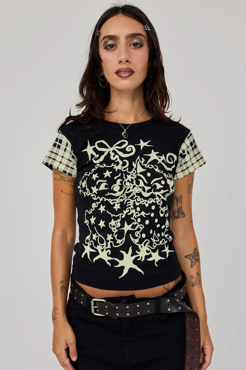 A woman modeling a black short-sleeve t-shirt with a cream-colored gothic-style graphic print of stars, swirls, and ornate flourishes. The sleeves feature a beige and black gingham check pattern.
