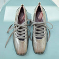 A pair of metallic taupe-brown lace-up shoes with a glossy finish, laid flat on a light blue textured surface.