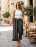 A woman walking on a cobblestone street wearing a white ribbed tank top and dark charcoal gray wide-leg linen pants, carrying a woven straw tote bag.