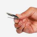 A close-up of a hand holding a small metallic clip with a green and silver patterned top, shown against a plain light gray background.