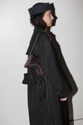 Person wearing a long black coat and dark hat with a brown bag, standing against a white wall.