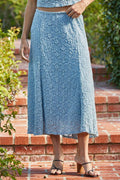 Person wearing a blue textured skirt standing on a brick path with greenery.