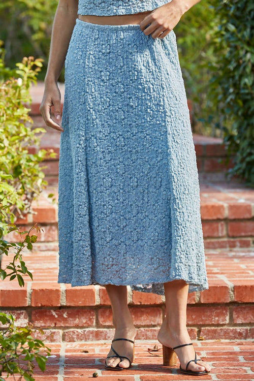 Person wearing a blue textured skirt standing on a brick path with greenery.