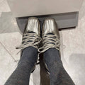 A pair of metallic silver lace-up shoes worn with dark gray socks, shown from a top-down perspective on a light gray tiled floor.