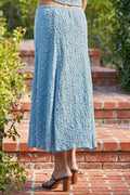 Person wearing a blue textured skirt standing on a brick path with greenery in the background