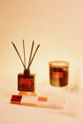 A diffuser and a candle on a white surface with a warm glow.