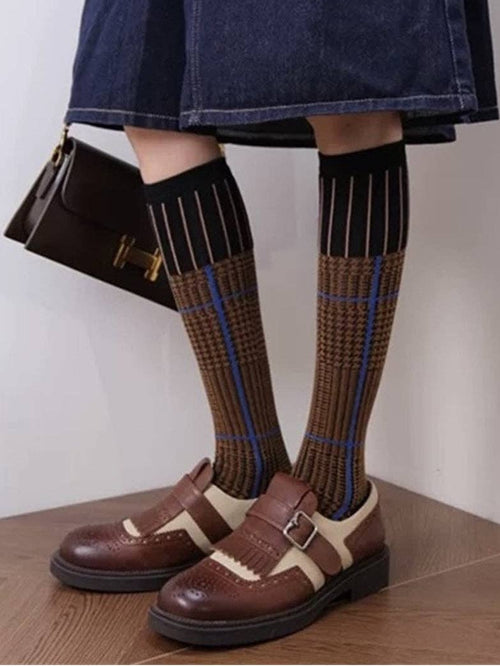 A person wearing knee-high brown patterned socks with black and blue stripes, paired with brown Mary Jane shoes and a dark denim skirt, holding a small black handbag.
