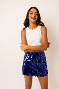 A woman modeling a white sleeveless top and a royal blue sequined mini skirt against a plain off-white background.