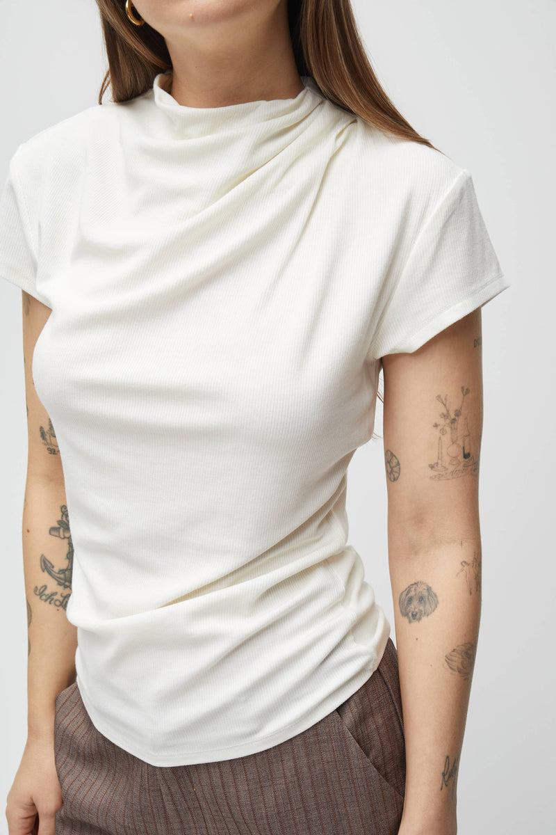 A close-up of a model wearing a white short-sleeve t-shirt with a draped, asymmetrical neckline and gathered waist details, paired with brown plaid trousers against a light gray background.