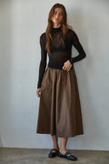 Woman wearing a black long-sleeve top and brown skirt against a plain background