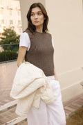 Woman wearing a brown knitted sleeveless top and white pants, holding a white jacket outdoors.