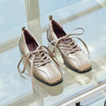 A pair of metallic rose gold low-top lace-up shoes with a glossy finish, displayed on a glass surface.